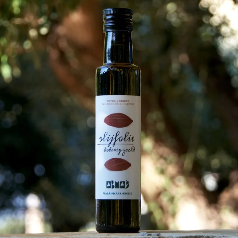 Fles Abma's olijfolie Boterig zacht buiten in de zon, met olijfbomen op de achtergrond