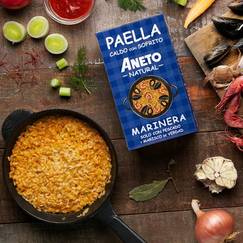 Pan met paella marinera naast een pak Aneto bouillon, tussen verse groenten en kruiden